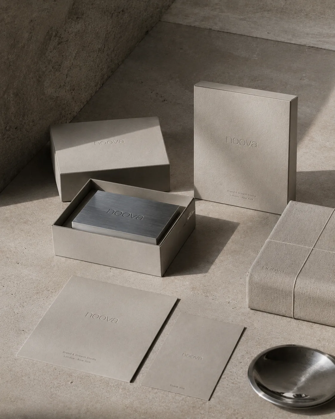 Warm grey packaging suite with embossed wordmarks and a brushed steel card box in soft light.